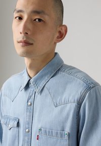 Lichtblauw denim shirt met puntige westerse yoke, zilveren drukknoopjes en twee opgestikte zakken aan de voorkant met groene accenten. Soepele textuur.