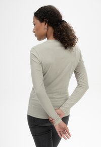 Langärmliges, hellgraues Shirt mit schmaler Passform, weichem Material und Rundhalsausschnitt. Das Modell steht mit den Händen hinter dem Rücken und trägt dunkle Jeans.