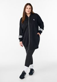 Melna uzvilkama kapuces džemperis ar baltām līnijām uz piedurknēm, sudraba logotipu, kombinēts ar melnām legingām un zamšādas sporta apaviem.