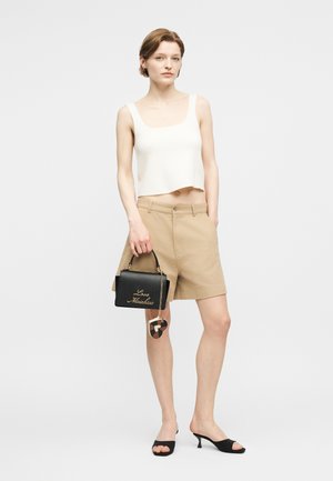 Donna con top corto bianco senza maniche e pantaloncini beige, che tiene una borsa nera con scritte dorate e un ciondolo decorativo, indossa tacchi neri.