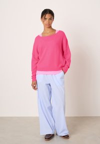 Pull oversize fuchsia avec un ourlet côtelé, superposé sur un haut rose clair. Un pantalon large rayé bleu pâle et des chaussures marron complètent la tenue.