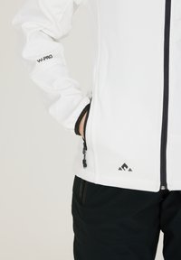 Vit softshell-jacka med dragkedjefickor, svarta accenter och logotyp. Har en hög krage och figursydda manschetter för en strömlinjeformad look.