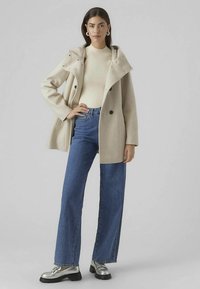 Beige wollen mix jas met capuchon, dubbele knopenrij en grote knopen, gecombineerd met lichtblauwe jeans met wijde pijpen en zilveren plateauzolen.