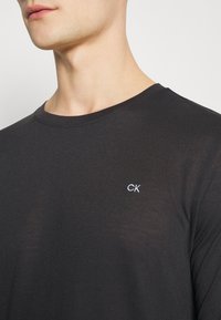 Black long-sleeve t-shirt made of soft fabric, featuring a round neckline and white embroidered "CK" logo on the left chest area.