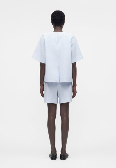 Άτομο με κοντά σγουρά μαλλιά που φοράει ανοιχτό μπλε υφασμάτινο κοντομάνικο τοπ και αντίστοιχα σορτς, στέκεται γυρισμένο προς τα πίσω σε απλό λευκό φόντο.