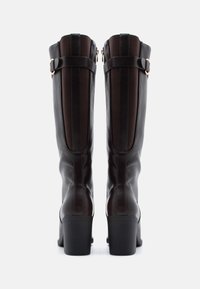 Botas altas de cuero marrón con un acabado liso y un ligero brillo. Presentan paneles de tela oscura y accesorios de hebilla en la parte superior. Diseño con tacón cuadrado.