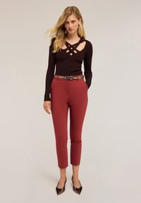 Maglione a coste marrone scuro con dettaglio a taglio, abbinato a pantaloni rossi aderenti e una cintura marrone chiaro. Tacchi neri a punta completano il look.