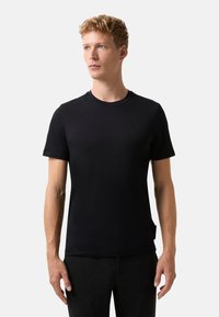 Młody mężczyzna z krótkimi, kręconymi blond włosami, ubrany w zwyk�ły czarny t-shirt z krótkim rękawem i ciemne spodnie, stojący na tle białym.