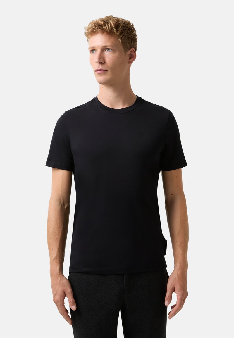 Młody mężczyzna z krótkimi, kręconymi blond włosami, ubrany w zwykły czarny t-shirt z krótkim rękawem i ciemne spodnie, stojący na tle białym.