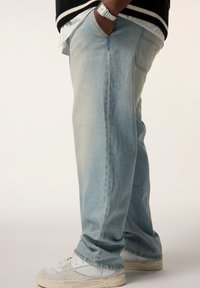 Helle blaue Denim-Jeans mit entspannter Passform, sichtbar mit Falten und Nähten; kombiniert mit weißen und grauen Sneakers.