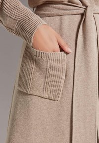 Beiger Cardigan mit gerippten Akzenten, einem Bindegürtel und großen aufgesetzten Taschen. Die Textur ist weich mit einem subtilen gewebten Muster.