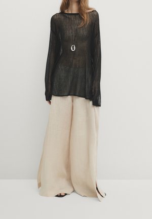 Woman wearing a sheer black long-sleeve top, beige wide-leg pants, a long pendant necklace, and black open-toe shoes.