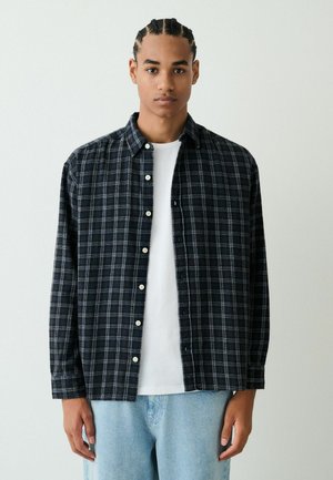 Schwarz-blau kariertes Flanellhemd mit Knopfleiste und weißen Knöpfen, über einem weißen T-Shirt getragen, kombiniert mit hellblauen Jeans.