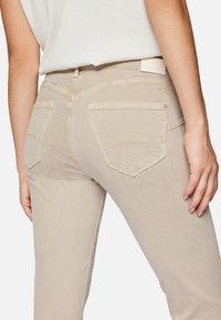Light beige jeans with a fitted design, rear pockets featuring curved stitching, and a smooth texture. Waist label visible at the back.
