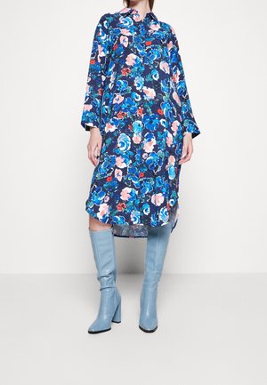Blumenmusterkleid in Marineblau mit blauen, pinken und roten Mustern; lange Ärmel und ein Kragen. Kombiniert mit hellblauen, kniehohen, Absatzstiefeln.