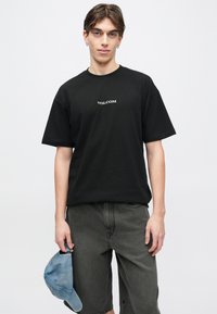 Νέος άνδρας με μαύρο t-shirt Volcom και σκούρα γκρι σορτς κρατάει ένα ανοιχτό μπλε καπέλο στο αριστερό του χέρι, σε λευκό φόντο.
