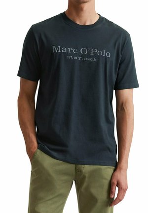 Uomo che indossa una t-shirt nera Marc O’Polo con la scritta "EST. IN STOCKHOLM", abbinata a pantaloni verde oliva, con le mani parzialmente nelle tasche.