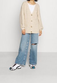 Béžový pletený kardigan s výstřihem do V, pěti velkými knoflíky a našitými rukávy, zkombinovaný se světle modrými širokými džínami s dírou a sportovními botami.