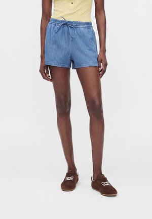 Person iført blå snørede shorts, gul ærmeløs top og brune sko, stående mod en ensfarvet hvid baggrund.