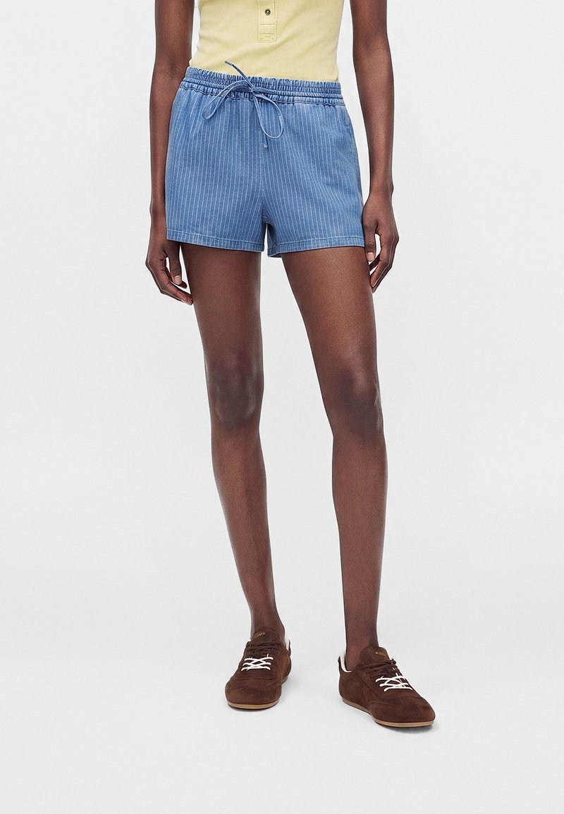 Personne portant un short bleu à cordon, un haut sans manches jaune et des chaussures marron, debout devant un fond blanc uni.