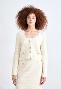 Elfenben struktureret cardigan med et cropped design, forsynet med guldfarvede knapper og hængsler som detalje, parret med en matchende nederdel.