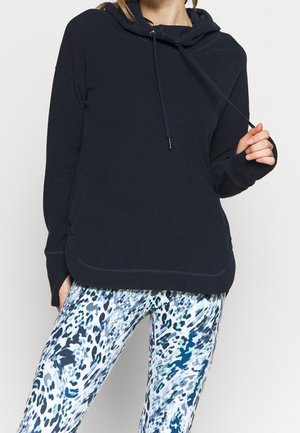 Frau trägt schwarzen Hoodie mit Daumenlöchern und blau-weiß-schwarz abstrakt gemusterte Leggings, hält einen Kordelzug.