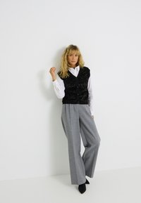 Gilet noir orné de sequins sur une chemise blanche à col, associé à un pantalon large gris et des chaussures à talons pointus noires, sur fond blanc.