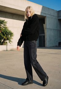 Mladý člověk s blond vlasy nosící černou fleecovou bundu, tmavé široké džíny a černé boty stojící venku poblíž betonových zdí.