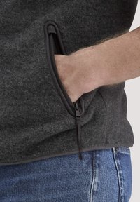 Graue Fleece-Weste mit einer Reißverschlusstasche. Der Stoff ist weich und strukturiert, mit einem kontrastierenden dunklen Reißverschluss und Saum. Getragen über blauen Jeans.