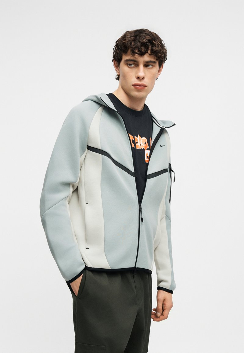 Junger Mann trägt einen hellgrauen und weißen Kapuzenpullover mit Reißverschluss über einem schwarzen T-Shirt mit Grafik, die Hände in den dunklen Hosentaschen, schaut zur Seite.