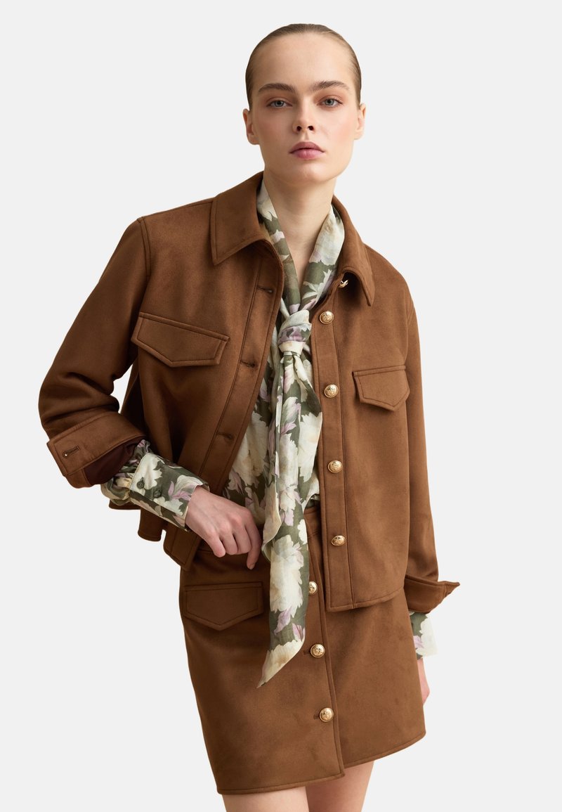 Mujer con chaqueta marrón abotonada y falda a juego sobre una blusa con pañuelo floral, posando con una mano en la cadera sobre un fondo liso.