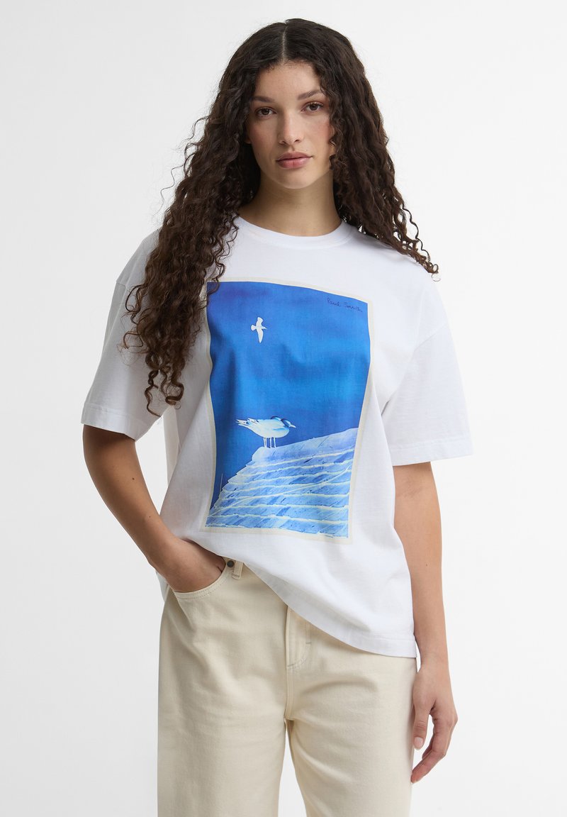 Γυναίκα με μακριά σγουρά μαλλιά φοράει λευκό μπλουζάκι με φωτογραφία της θάλασσας, γλάρους πάνω σε βράχια κάτω από έναν φωτεινό γαλάζιο ουρανό.