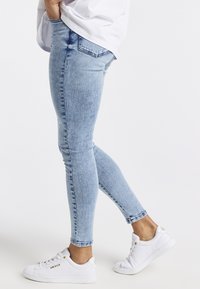 Persona vestida con jeans ajustados de un azul claro y zapatillas blancas con letras doradas, de pie con una pierna ligeramente flexionada.