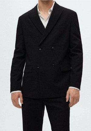 Homme portant une veste de costume noire croisée sur une chemise blanche, debout devant un fond clair uni, cadré au niveau de la tête et des genoux.