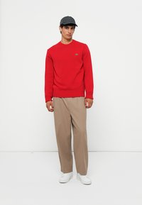 Pulover roșu tricotat cu tiv împletit, pantaloni largi bej și adidași albi. Modelul poartă o șapcă închisă, stând în fața unui fundal alb.
