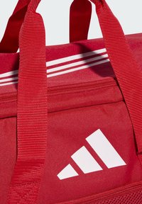 Borsa duffel rossa con tessuto testurizzato, presenta un design a tre strisce bianche e il logo. Manici robusti con un motivo rinforzato.