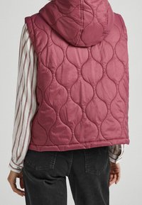 Gestepptes Vest in tiefrosa Farbe mit Kapuze. Verfügt über gerundete Muster und eine lässige Passform, kombiniert mit einem gestreiften Langarmshirt.