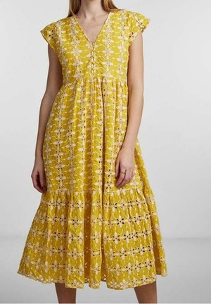 Kobieta ubrana w żółtą sukienkę midi z białą kwiatową haftem i falbanowymi rękawami, stojąca na jednolitym tle.