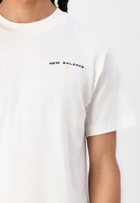 Fehér pamut póló lekerekített nyakkivágással és rövid ujjakkal. A mellkason feketével hímzett "NEW BALANCE" logó sárga akcentussal.