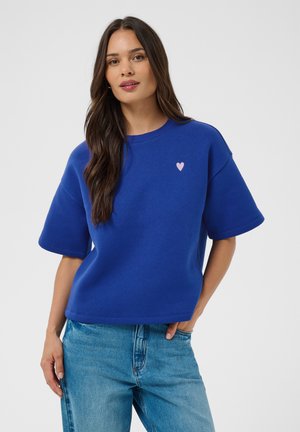 Femme aux longs cheveux foncés portant un pull bleu à manches courtes avec un petit motif cœur et un jean bleu clair, debout devant un fond uni.