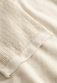 Detailný záber na krémovo sfarbenú pletenú látku s rebrovaným nápletom, ktorý má štruktúrovaný, vzorovaný pletený dizajn.