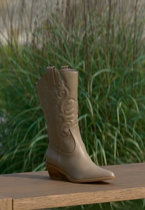 Lysebrun lær cowboystøvel med brodert design og tresåle plassert på en treflate mot grønn plantebakgrunn.