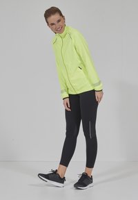 Hellgelbe, leichte Jacke mit Reißverschluss, langen Ärmeln und reflektierenden Akzenten, kombiniert mit schwarzen Leggings und Sportschuhen.