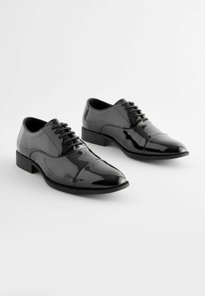 Scarpe da abito in pelle verniciata nera con punta affilata, design allacciato e tacco basso. Superficie liscia con una leggera lucentezza.