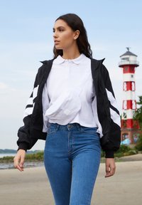 Noor naine kandmas valget polot, musta jopet, siniseid teksapükse ja valget õlakotti, kõndimas punase-valge majaka lähedal ranna ääres.