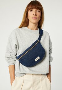 Sac banane en tissu navy à la forme courbée, avec une fermeture éclair argentée et une étiquette blanche. Porté en travers du corps sur un sweat-shirt gris clair.