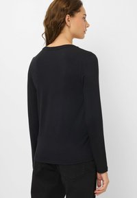 Donna con capelli castani che indossa una maglietta nera a maniche lunghe e pantaloni neri, vista di profilo su sfondo bianco.