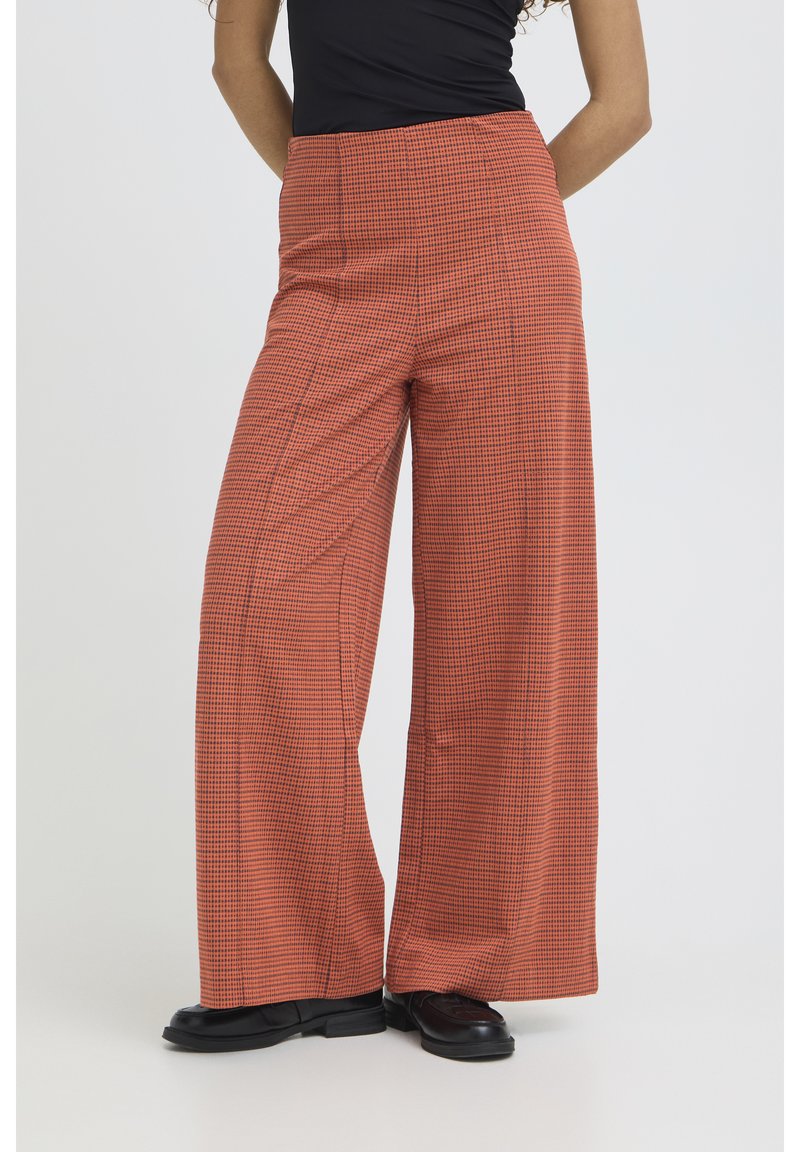 Persona con pantalones anchos a cuadros naranjas y negros, zapatos negros y una camiseta negra, de pie frente a un fondo liso.