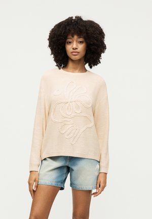 Γυναίκα με μπούκλες φοράει ένα μπεζ πουλόβερ με ανάγλυφο floral σχέδιο και ανοικτό μπλε τζιν σορτς, στέκεται μπροστά σε απλό φόντο.