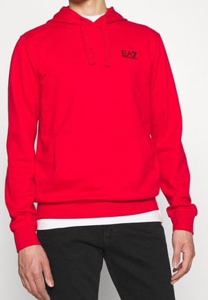 Mann trägt roten EA7 Emporio Armani Kapuzenpullover mit Fronttasche, Kordelzugkapuze, weißem Unterhemd und schwarzer Hose vor schlichte Hintergrund.